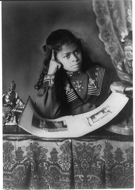 African American girl, half-length portrait, with right hand to cheek, with illustrated book on table
